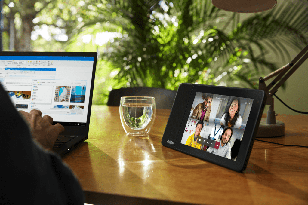 WFH seen with a large plant in the background, a person's hands on a laptop and the ThinkSmart View set up to their left with a video meeting onscreen.